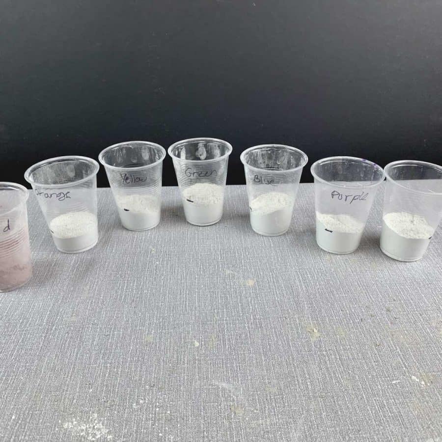 Concrete Candle Holder- 7 plastic cups lined up with concrete mix filled to 2 oz line. Each has name of rainbow color on them.