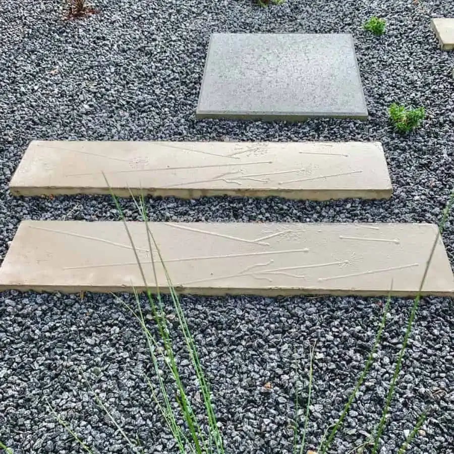 two rectangular concrete pavers with protruding details of dandelions and other pretty designs, laying on a gravel pathway