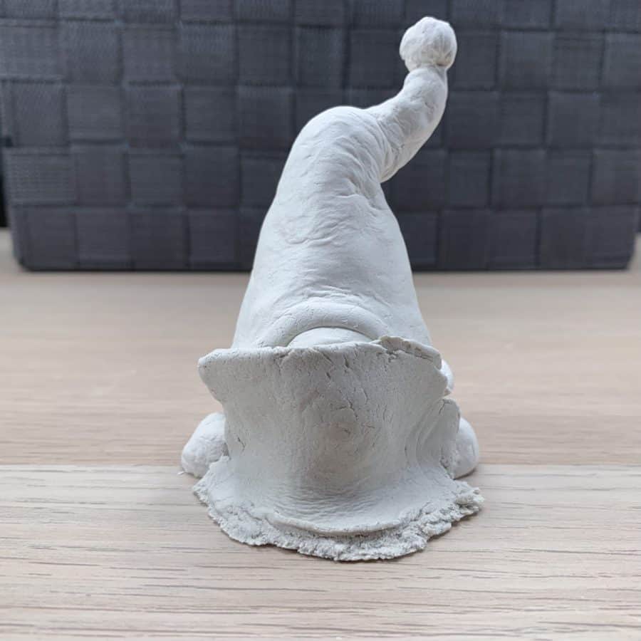 air dry clay gnome standing on feet with piece of rectangular shaped clay across his face and flared at bottom where it meets the table.