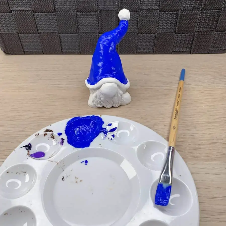 a clay gnome with the hat painted blue and a paint tray with a paintbrush on it.