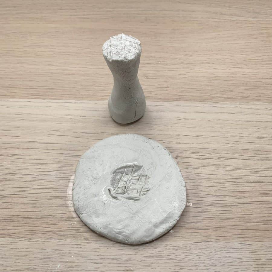 flat pancake shaped clay with center carved out and hatch marks, larger than first cap piece. Next to tall stem piece with hatch marks on top.