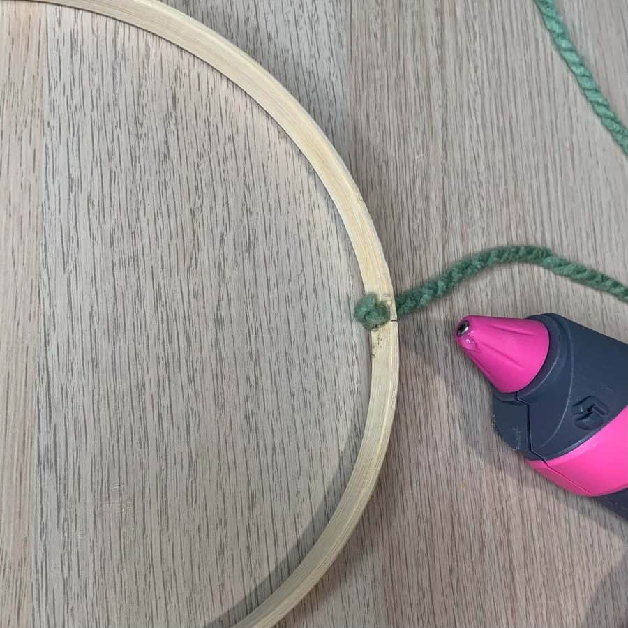tip of hot glue gun next to embroidery hoop with end of yarn glued inside
