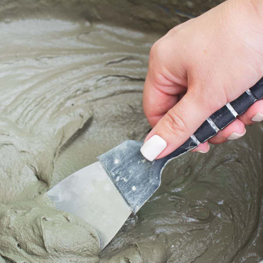A hand scooping wet cement mix with a spatula from a large bin.