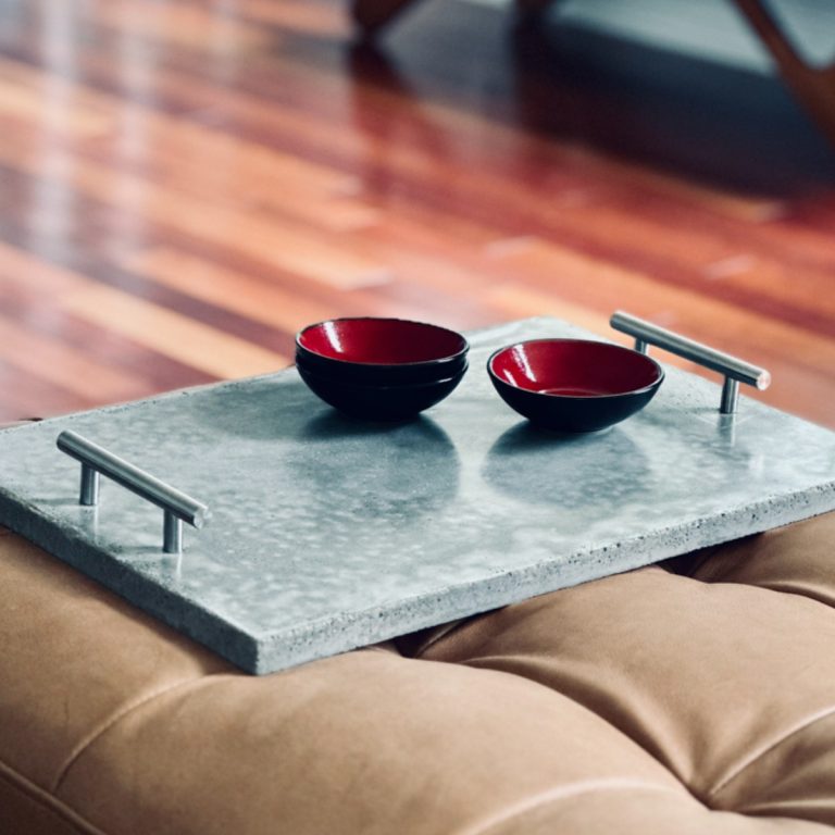 A smooth-looking concrete tray with handles on top of a leather ottoman. The tray has contrasting red dishes on it.