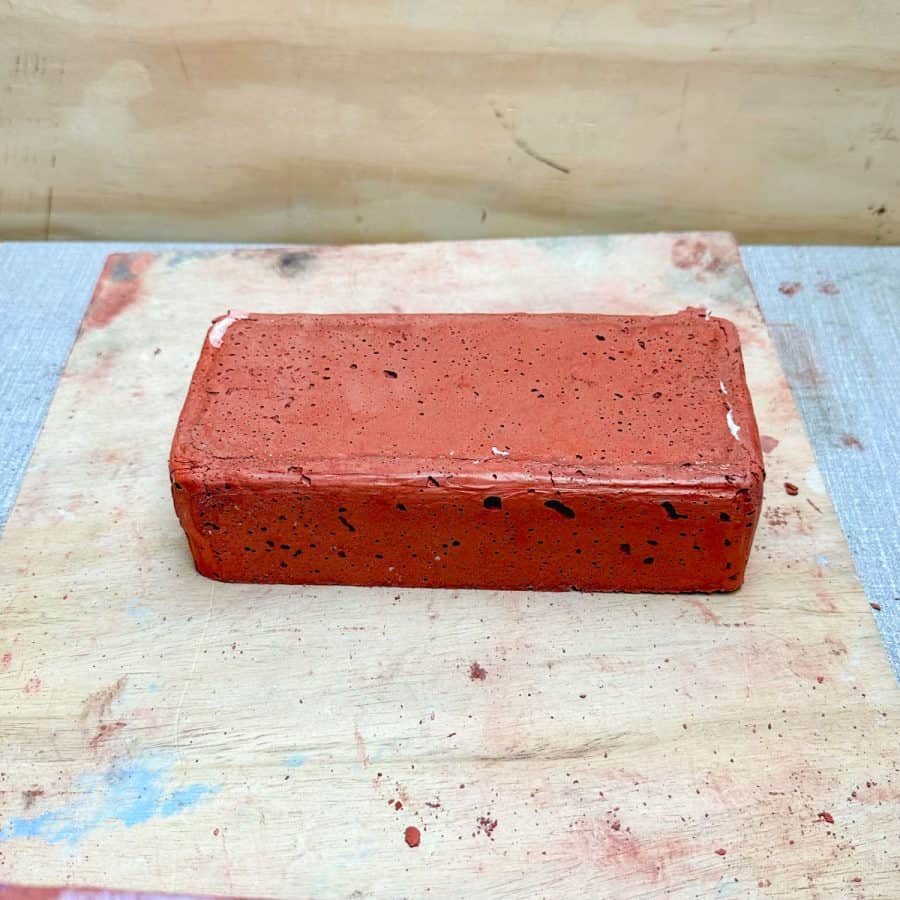 Brick red concrete brick on a table. Has rounded edges and a little bit of silicone caulk stuck between some folds.