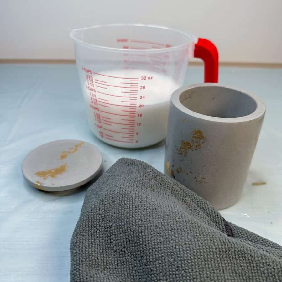 A plastic container with sealer in it and a concrete candle jar and lid and cloth next to it.