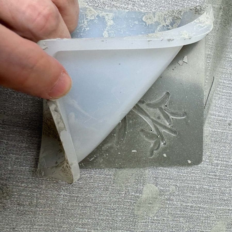 A hand peeling back a tile mold revealing a silicone stamp embedded into cured concrete.