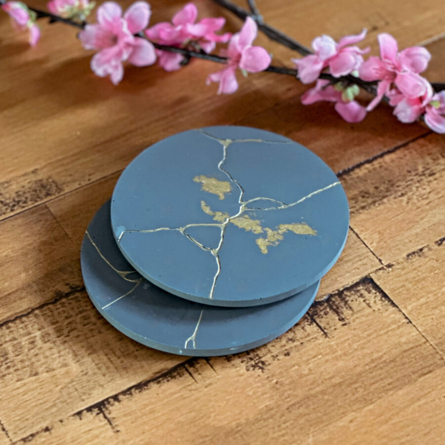 Two stacked Jesin kintsugi coasters with gold lines running through them that are handmade. They are on a table with flowers behind.